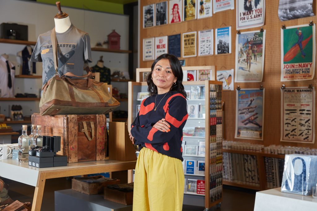 Portrait of Reasey Kheng, Buying Manager at Imperial War Museums. Reasey is pictured in one of the museum’s main shops, surrounded by products such as posters, a bag and a t-shirt.