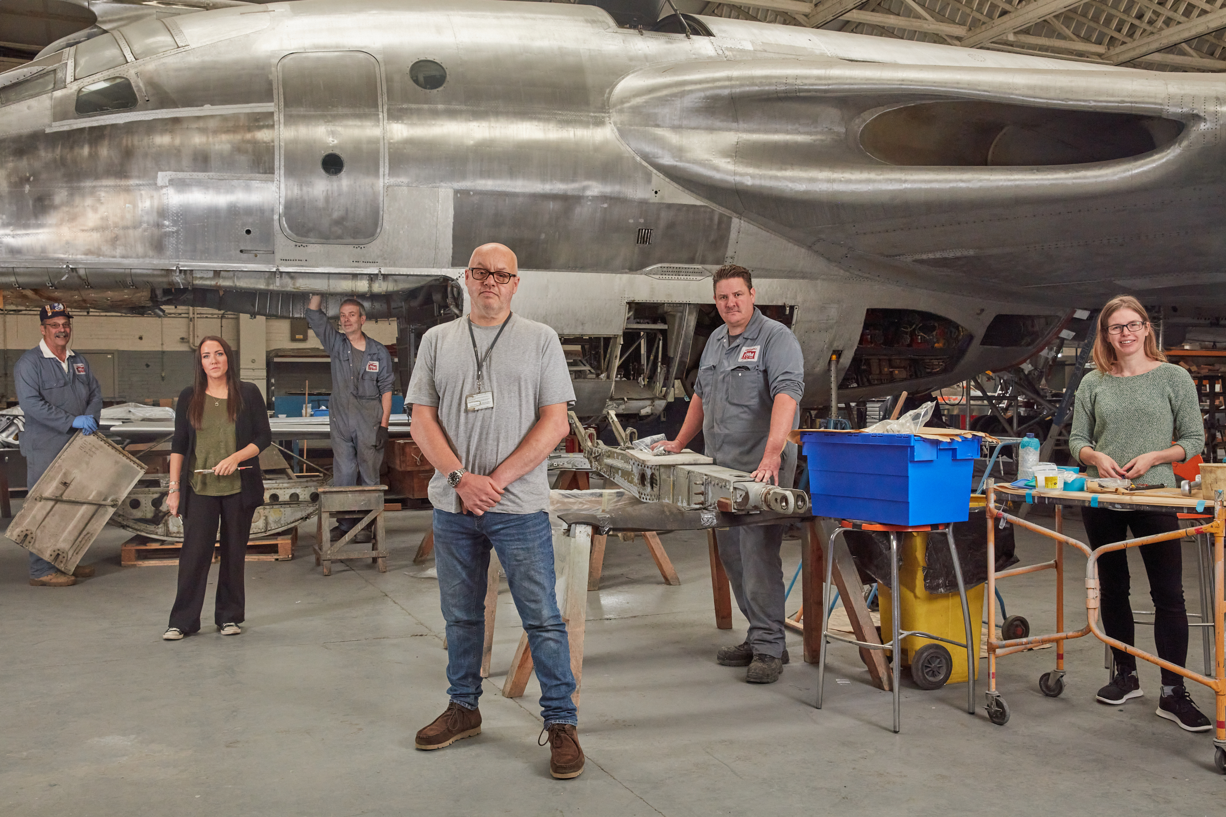 Portrait of the conservation team at IWM Duxford, UK. Six members of the team are pictured in an aircraft hanger in front of a silver aeroplane that they’ve been working on.