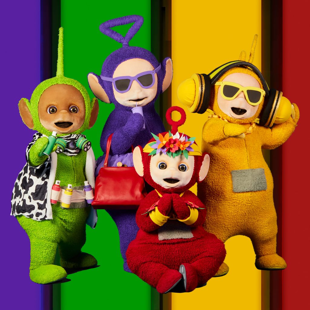 Portrait of the Teletubbies against a purple, green, yellow and red striped background. Dipsy is wearing a cow print jacket and holding some cans of spray paint. Tinky winky has sunglasses on and is sipping on a coffee. Laa-Laa has headphones as well as a gold chain on and is DJing. Po's head is adorned with flowers and is doing a yoga pose. 