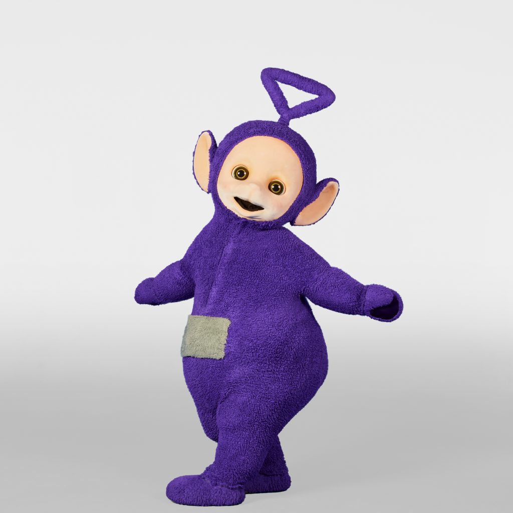 Tinky Winky is posing in a white studio with both hands out to the side.