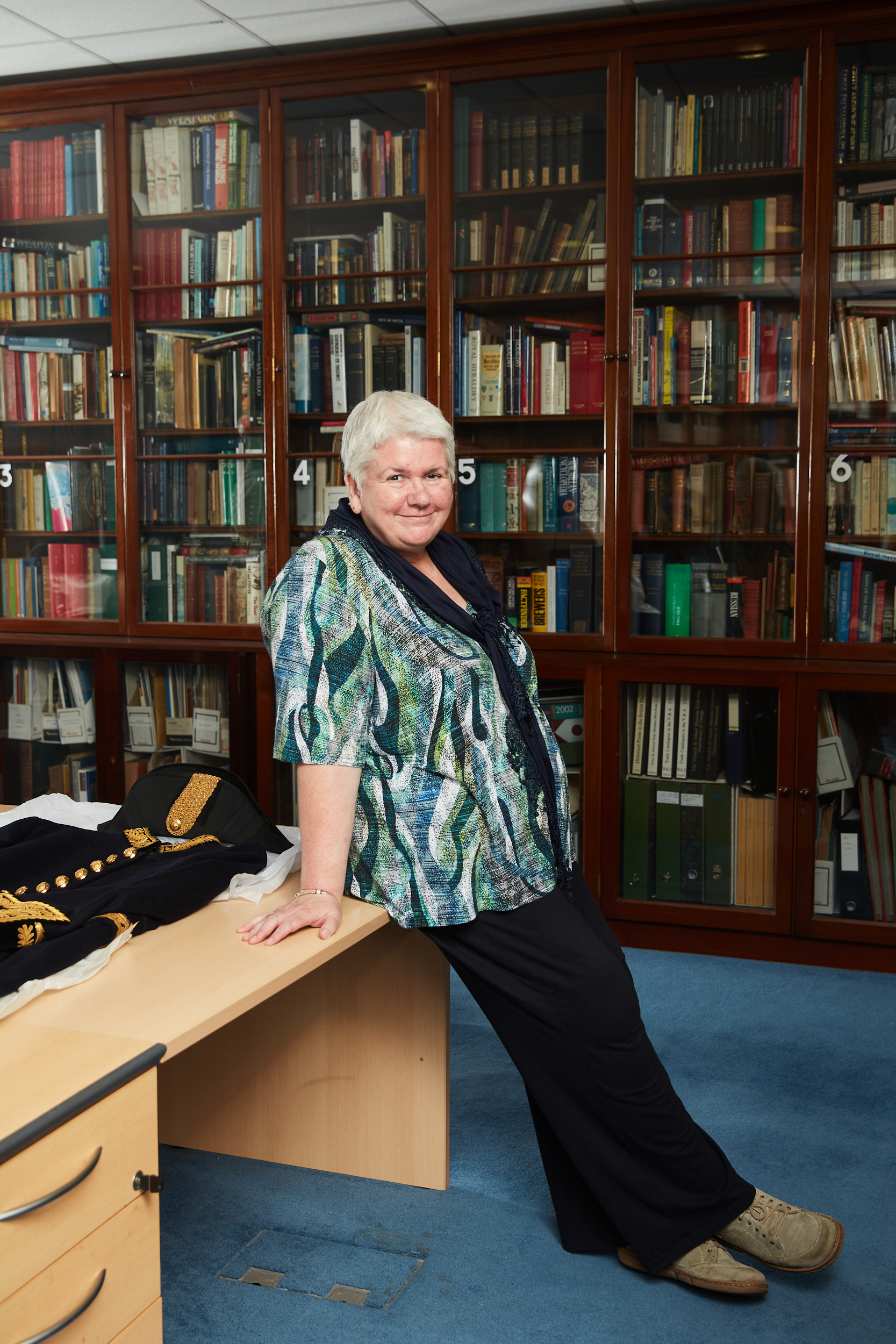 Bridget Clifford, keeper of Tower Armouries at the Tower of London, photographed leaning on a table. She is pictured smiling and standing in a room full of books in glass cabinets, with a military uniform placed on the table she leans against.