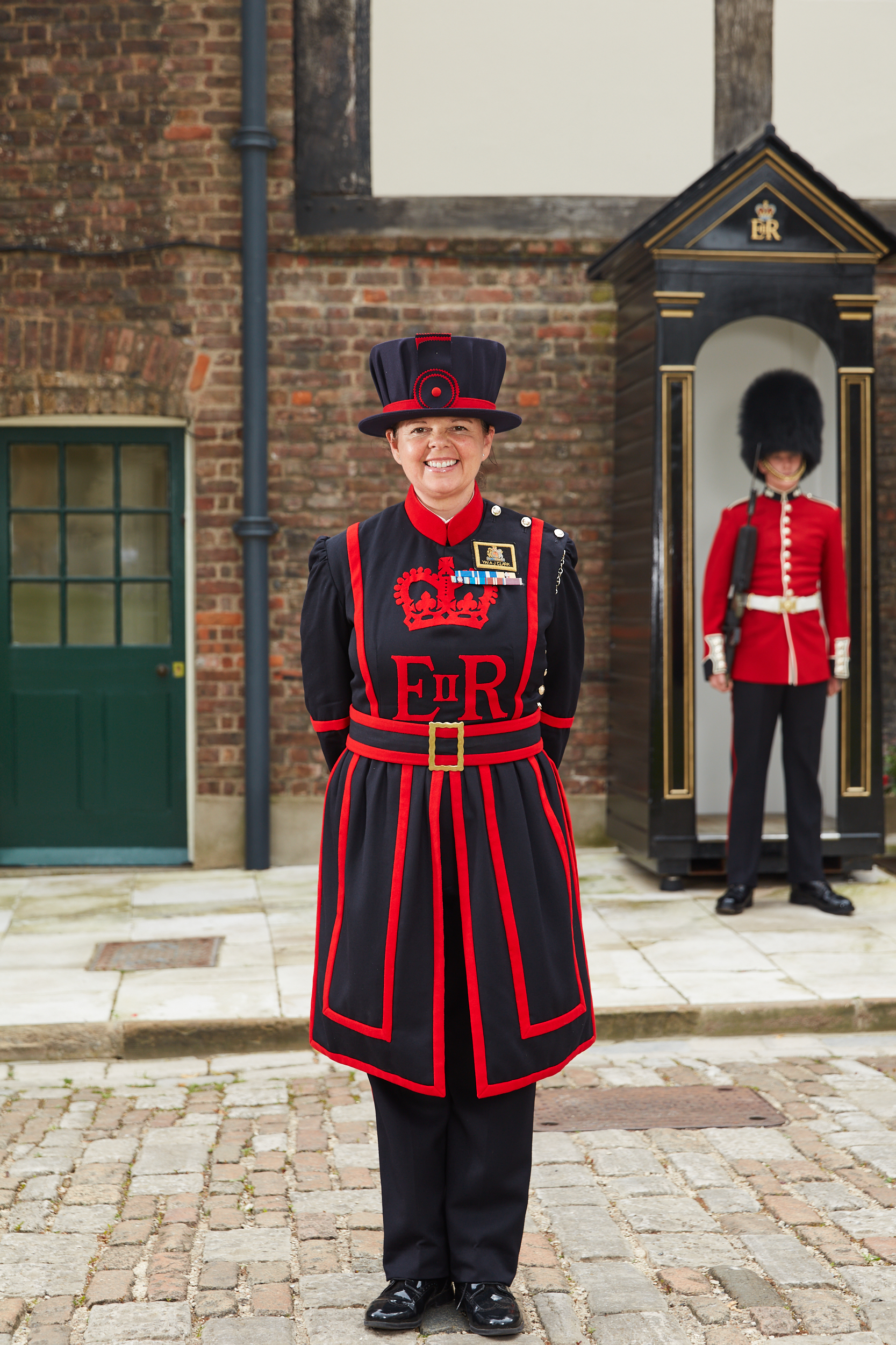 Portrait of beefeater AJ Clark. She is smiling with her hands behind her back within a Tower of London courtyard, and is wearing typical navy beefeater clothing. To her left stands a traditional royal guard. 