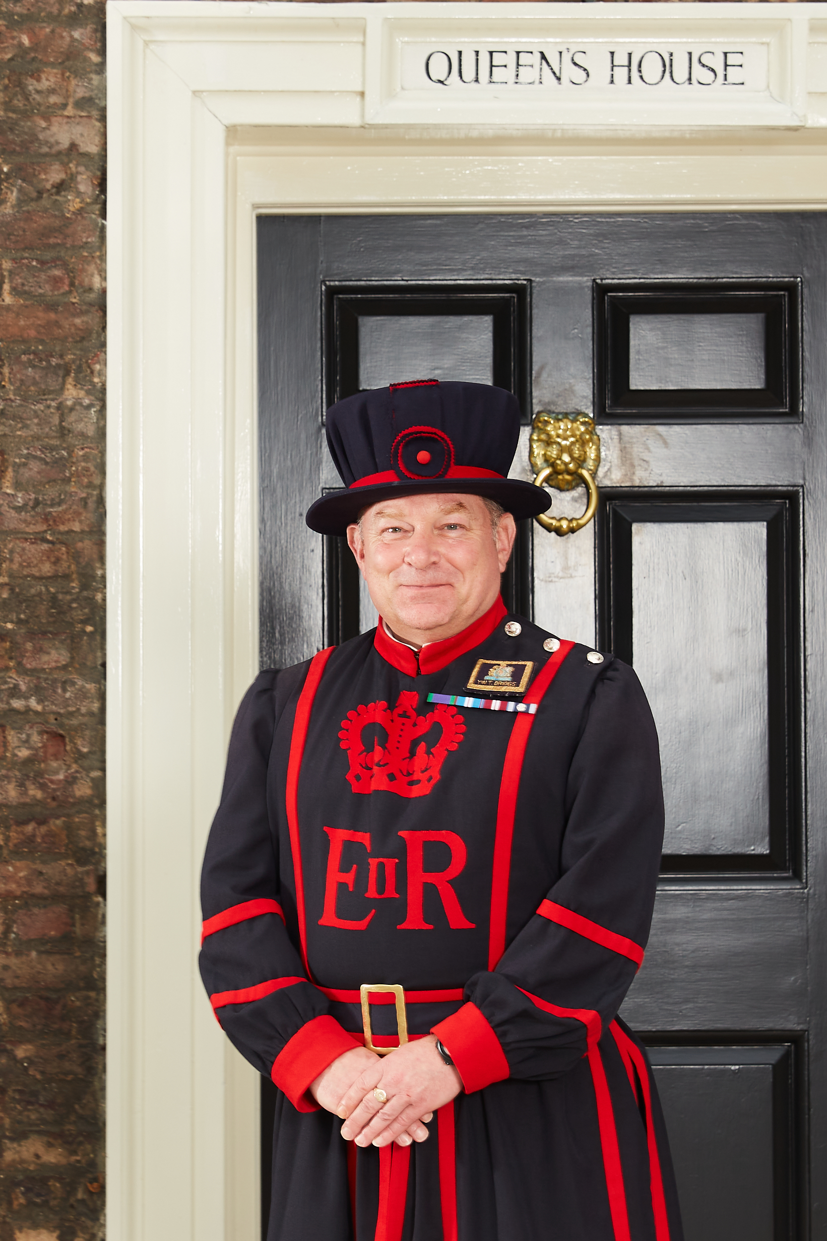 Beefeater Terry Briggs stands in front of a door labelled "Queen's Door" at the Tower of London. He is smiling with his hands held in front of him. 