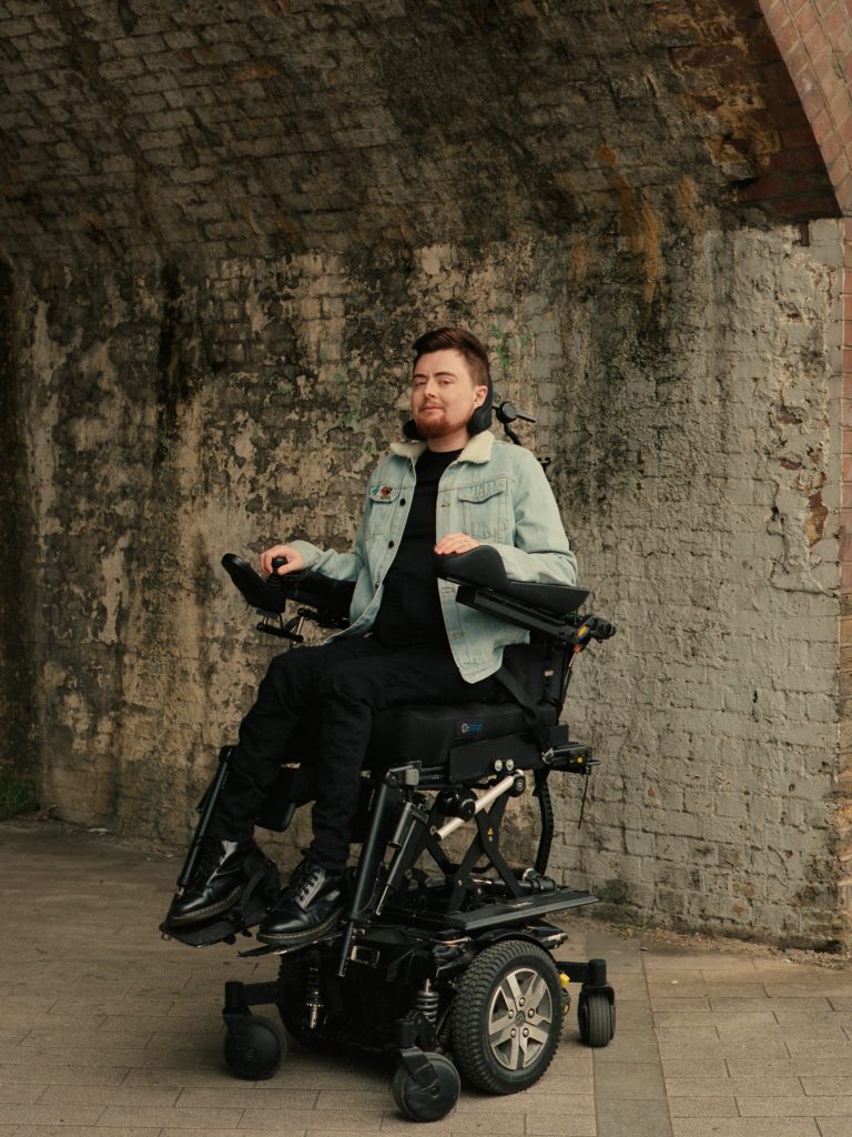 Jamie, a white person with auburn hair and an auburn beard is outside in their electric wheelchair in front of a textured stone arch. They are smiling, and turned slightly away from the camera but looking into the lens. They are wearing a pale blue denim jacket paired with a black top and black jeans. 