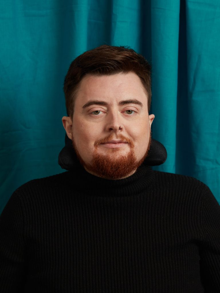 Jamie, a white person with auburn hair and an auburn beard is pictured smiling in front of a royal blue fabric background. They are wearing a black polo-neck top. The image is cropped from the chest up. They are slightly smiling, and looking at the camera. The headrest of their electric wheelchair can be seen behind their head.