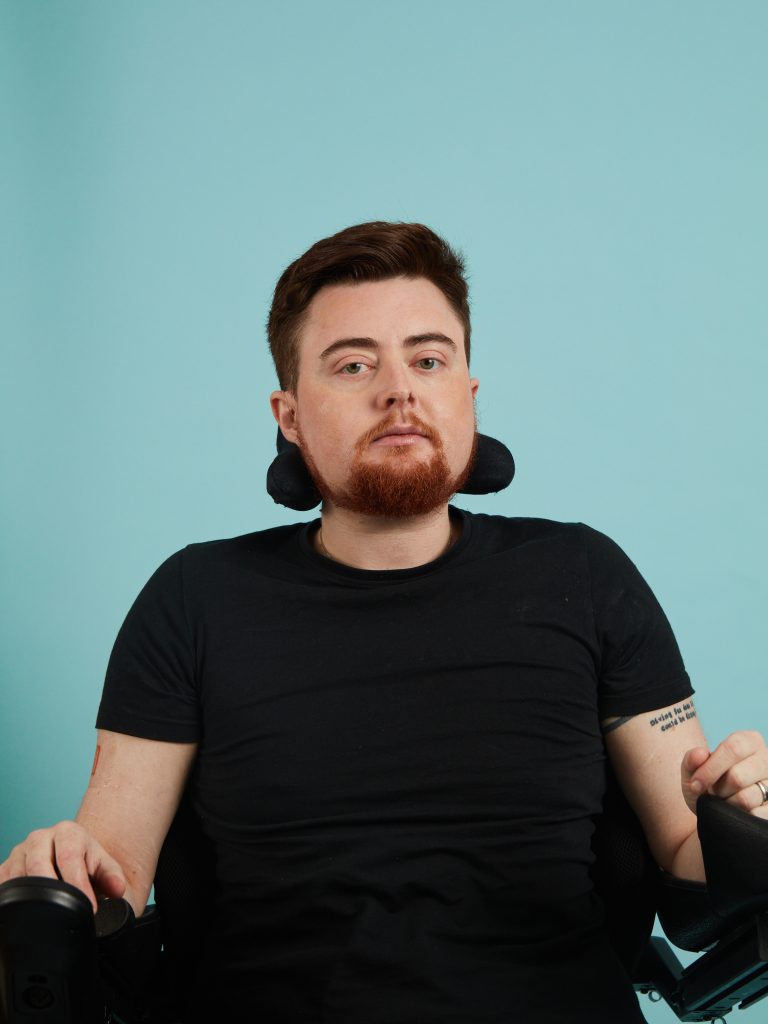 Jamie, a white person with auburn hair and an auburn beard is pictured at home in their electric wheelchair. They are wearing a black short-sleeved top. The image is cropped from the waist up, and they are in front of a pale blue photographic background. They have a thoughtful expression. 