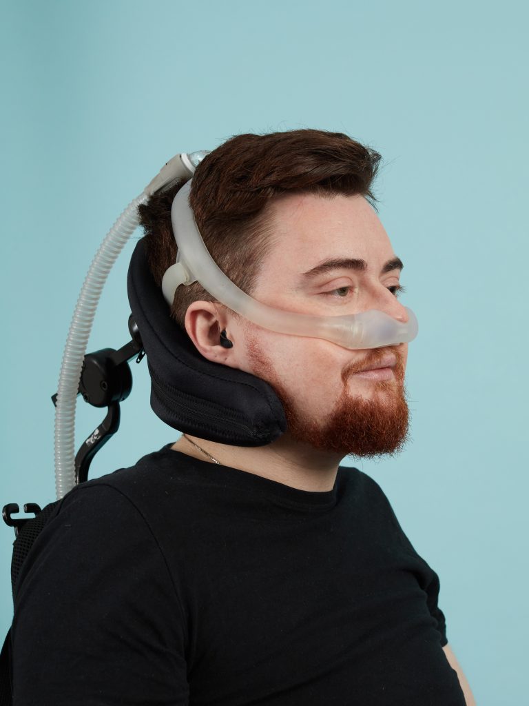 Jamie, a white person with auburn hair and an auburn beard is pictured smiling in a black top in front of a pale blue fabric background. They are wearing ventilation equipment and faced away from the camera. They are sat in their electric wheelchair, and their headrest is visible. The image is cropped from the shoulders up. 