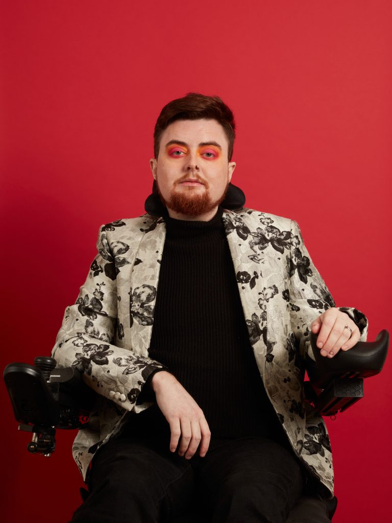 Jamie, a white person with auburn hair and an auburn beard, is sat in their electric wheelchair, with their right hand placed on their lap and their left arm on their armrest. They are looking directly into the camera. Jamie has bright orange and pink sunset eye make up on, and they are wearing a white jacket speckled with black flowers. The outfit is paired with a black shirt and black jeans. Behind them is a bright red background. 